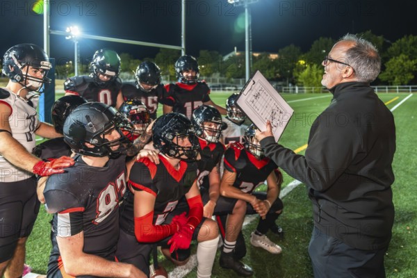 American football coach holds clipboard with play diagram, instructing young athletes in full uniform and helmets during a nighttime huddle on bright stadium turf field