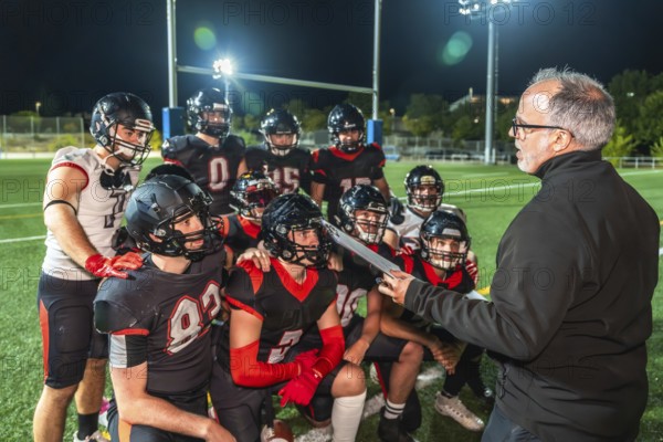 American football coach discussing strategy with his team of players in uniform and helmets during a nighttime practice or game on a green turf field under bright stadium lights