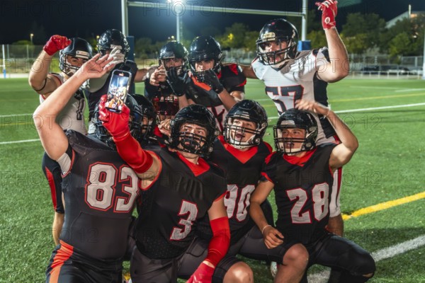 American football teammates in uniform celebrate a win on the field, smiling and posing for a group selfie as one player holds a smartphone under stadium lights