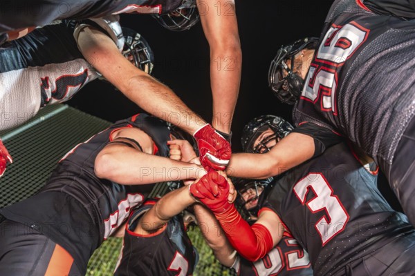 Football teammates in a night huddle, gloved fists stacked in the center, conveying unity, teamwork, power and focused strategy before the big play under stadium lights