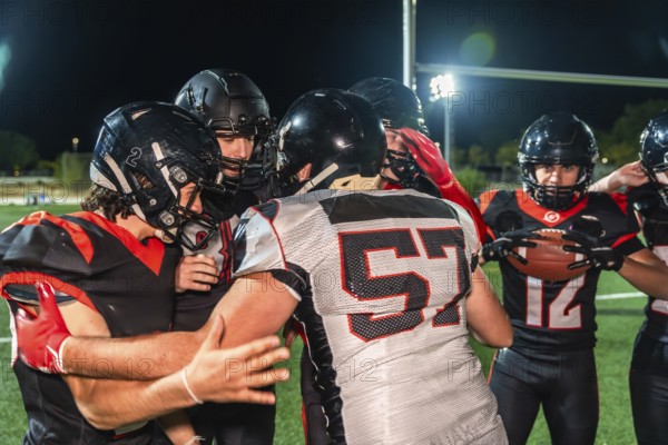 American football players in black and red uniforms with helmets and shoulder pads huddling on a green turf field during a night game, discussing strategy and demonstrating teamwork