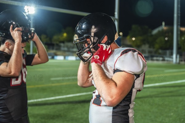 American football players adjusting their helmets on illuminated green field, preparing for intense training session or competitive game under the night sky
