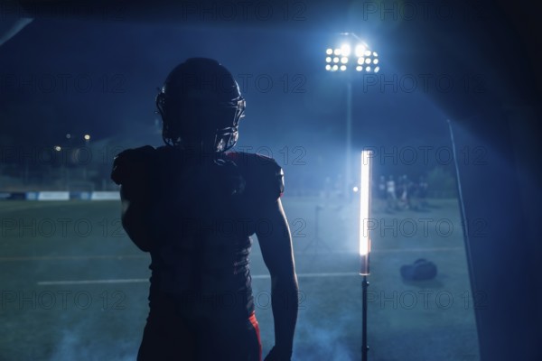 Football player standing in silhouette, ready to enter the stadium field at night, stadium lights creating a dramatic atmosphere, preparing for the game, showing determination