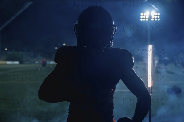 American football player standing in silhouette on a dark field under bright stadium lights at night, creating a dramatic atmosphere during an intense game or practice session
