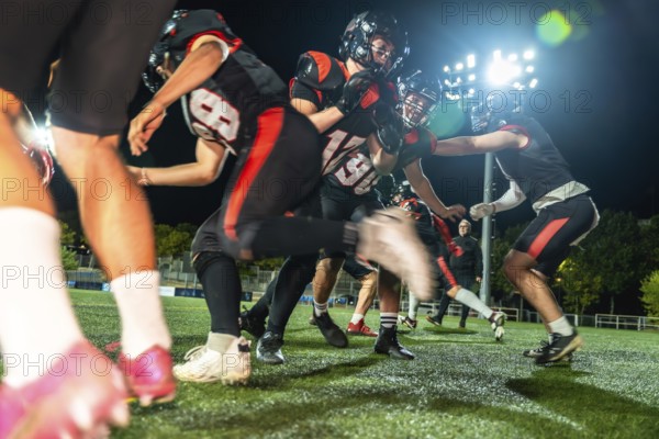 American football players in black and red uniform helmets battling on a green artificial turf field illuminated by stadium lights at night during a tackling play