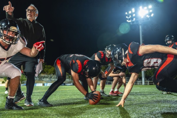 American football team huddled on a dark stadium field under bright lights at night, coach pointing and instructing players as they prepare for the next play and strategy
