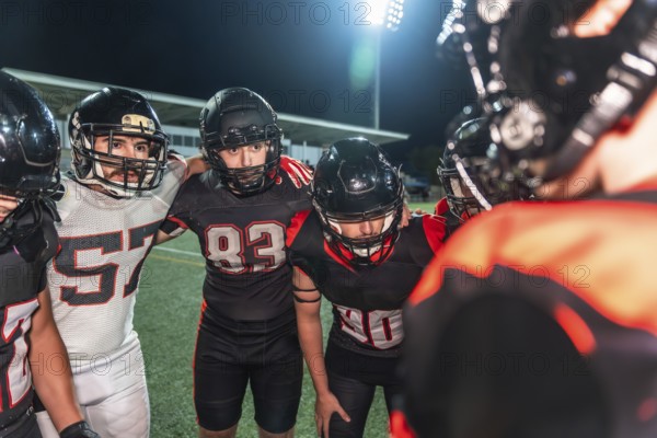 American football players in uniform and helmets huddling together with arms around shoulders, strategizing on a field under stadium lights at night during a game