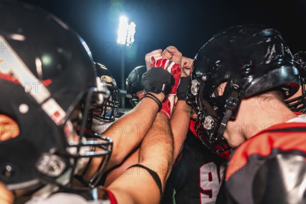 American football players putting hands together in a huddle, symbolizing teamwork, unity, strategy, and motivation on a field during a night game under stadium lights