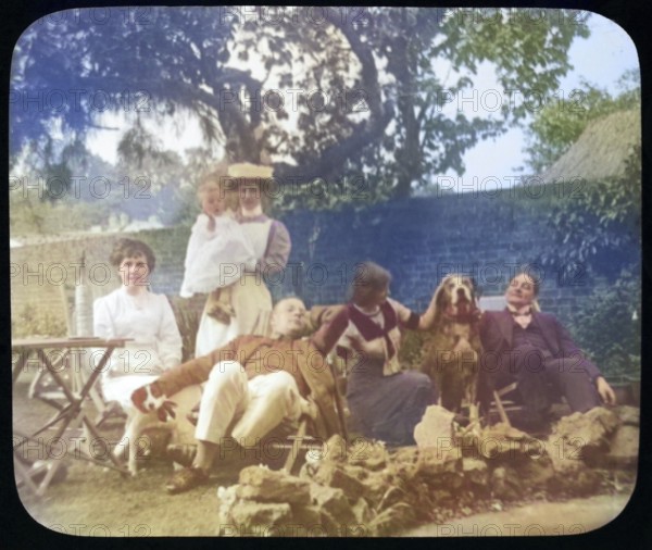 Affluent Edwardian family group men and women with nursemaid holding baby, outdoors in garden sitting in deckchairs with dogs, England, Uk, 1900 - 1930s colourised