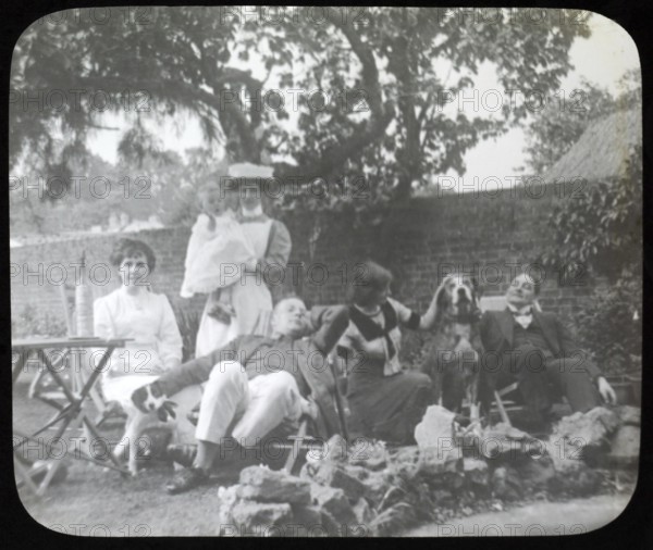 Affluent Edwardian family group men and women with nursemaid holding baby, outdoors in garden sitting in deckchairs with dogs, England, Uk, 1900 - 1930s