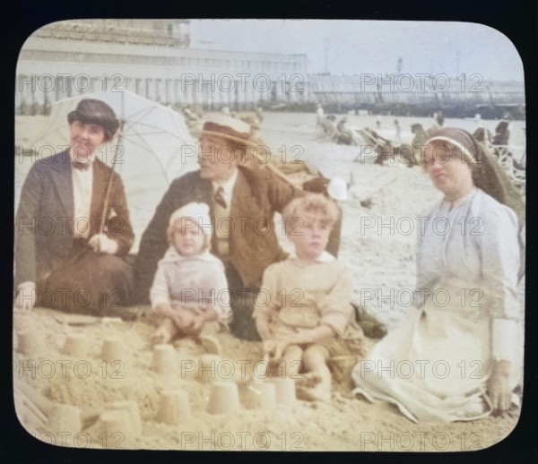 Affluent Edwardian family sitting on sandy seaside beach man, woman, children with nursemaid England, UK, 1900 - 1930s colourised