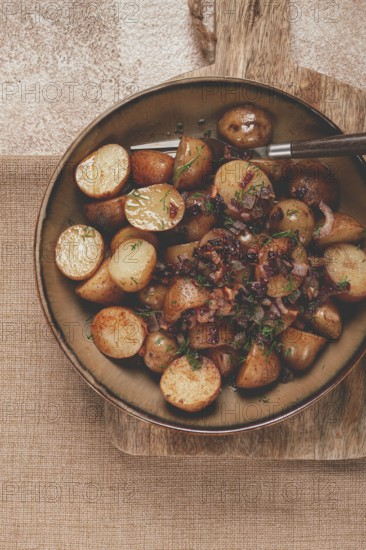 A bowl of perfectly roasted potatoes garnished with fresh herbs and caramelized onions sits on a wooden surface. The warm colors and rustic feel create an inviting atmosphere