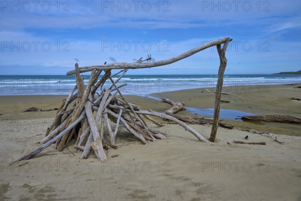 Treibholzskulptur Zelt am Strand unter blauem Himmel, ruhiges Meer im Hintergrund, Kaka Point, Clutha District, Otago, Südinsel, Neuseeland