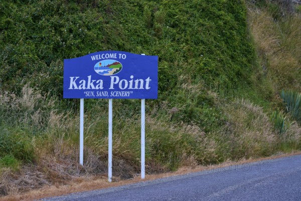 Willkommensschild von Kaka Point neben einer Straße mit grüner Vegetation und landschaftlicher Umgebung, Kaka Point, Clutha District, Otago, Südinsel, Neuseeland