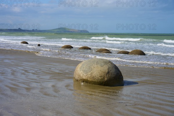 Große runde Felsen am Strand mit Wellen im Hintergrund und blauem Himmel, Moeraki Boulders, Moeraki, Hampden, North Otago, Südinsel, Neuseeland