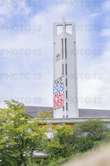 Church tower with protest banner between trees under blue sky, Tesla Model Y Juniper, Gechingen, Black Forest, Germany