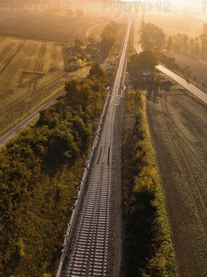 Aerial view of a landscape with railway tracks and fields in the warm autumn light of sunrise, a road follows the route of the tracks, Hermann Hesse Bahn, Ostelsheim, Black Forest, Germany