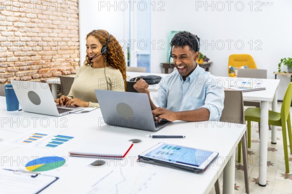 Diverse colleagues with headsets performing online customer service in an open plan office. A smiling woman typing on a laptop. And a man expressing excitement with a fist pump after achieving a goal