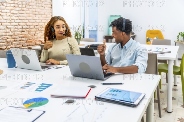 Diverse young professionals working in a modern office, wearing headsets, using laptops, and exchanging thumbs up gestures, symbolizing successful collaboration and positive customer service