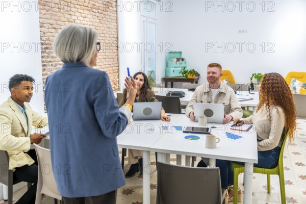 Senior professional woman presenting during a business meeting with a diverse group of young colleagues collaborating and discussing ideas in a modern office space