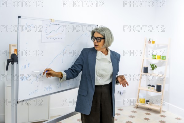 Senior businesswoman actively explaining financial data and market trends on a whiteboard during a presentation, sharing knowledge and expertise in a professional office setting