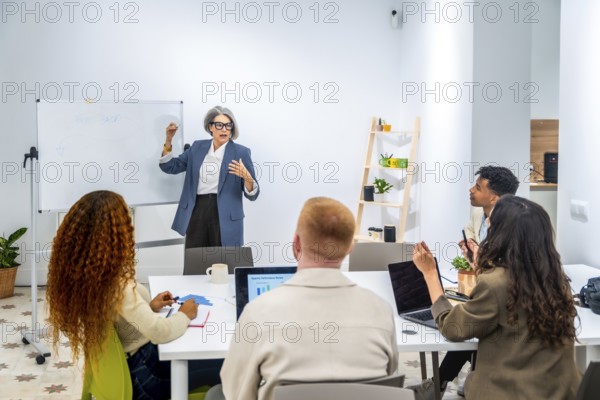 Senior grey haired business woman coaching and training a group of young, diverse professionals in a modern office, standing by a whiteboard and explaining strategy during a collaborative meeting