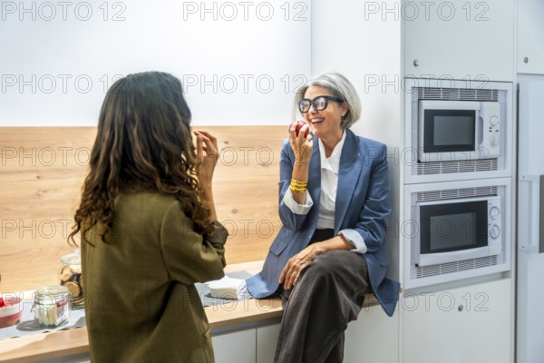 Two business women colleagues having a healthy snack, eating apples and laughing together while taking a break in the modern office kitchen or canteen area