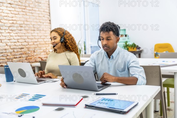 Multiracial customer service team wearing headsets and typing on laptops in a modern office, providing friendly tech support, helpline assistance and collaborative problem solving