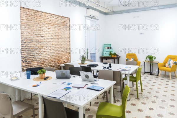 Bright open plan coworking office space featuring long white tables with laptops and charts, a textured brick wall, and comfortable yellow armchairs in a relaxed meeting corner