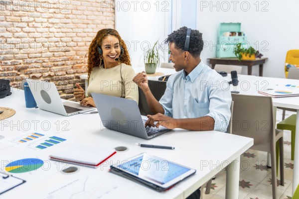 Two smiling multiracial coworkers wearing headsets, collaborating and working on laptops at a modern office desk, celebrating success and teamwork in a customer support environment
