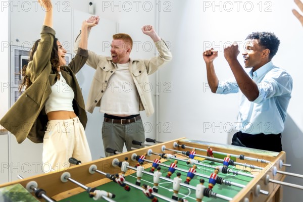 Diverse group of friends raising their arms in celebration after winning a foosball match, enjoying a fun and competitive break time in a modern office or coworking space