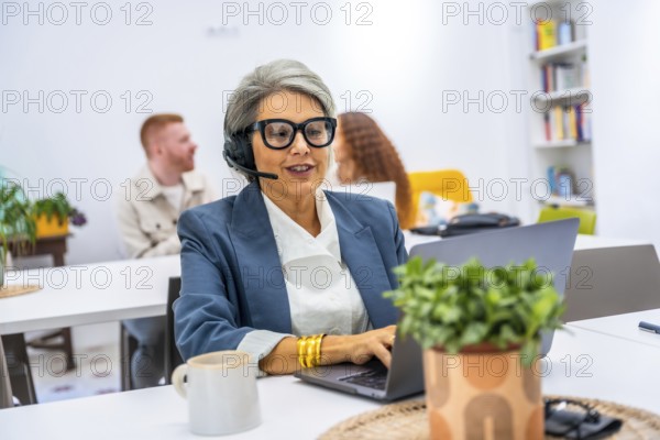 Senior woman actively working in a modern office, wearing a headset and spectacles while typing on a laptop, engaging in customer service or remote communication in a collaborative workspace
