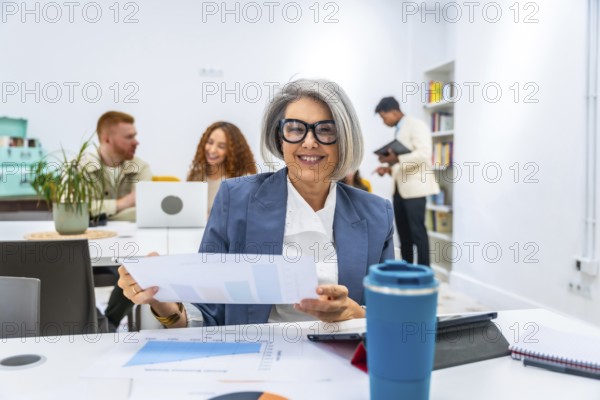 Happy mature businesswoman smiling at the camera, holding charts and graphs during a meeting with diverse colleagues collaborating in a dynamic corporate office environment
