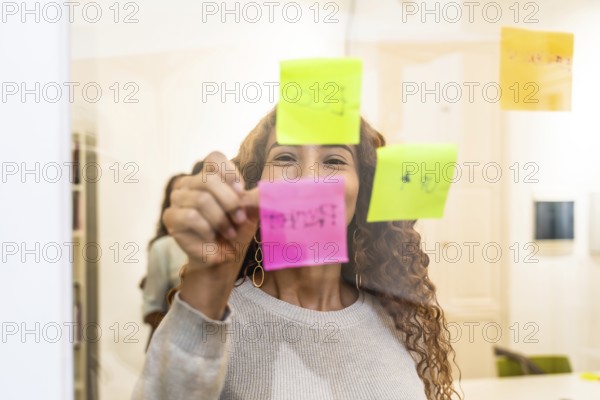 Happy woman collaborating on a project, writing ideas and tasks on colorful sticky notes on a clear glass wall in a modern office during a lively planning session