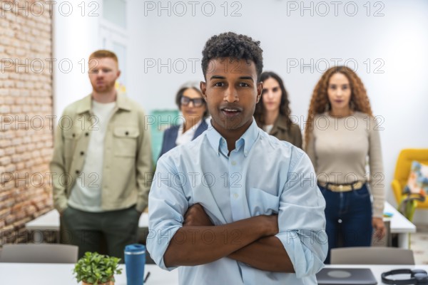 Young man standing with arms crossed, confidently looking at the camera, with his diverse team members blurred in the background, working together in a bright, contemporary office
