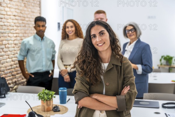 Confident young woman with curly hair smiling at camera, standing with arms crossed in a modern office, leading a diverse team of professionals in the background