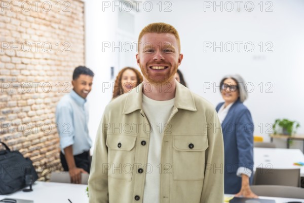 Redhead man standing in front, confidently smiling at the camera, with a diverse business team blurred in the background, working together in a modern office