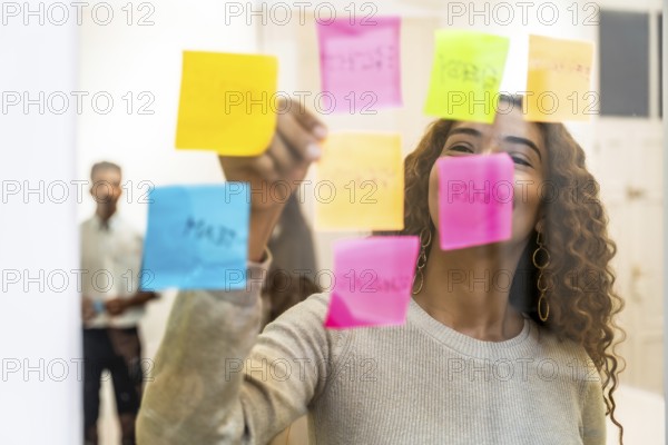 Woman organizing colorful sticky notes on a clear glass board, actively brainstorming and planning new business strategies in a modern office environment