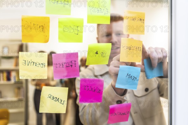 Man in a modern office space arranging colorful sticky notes with various concepts and keywords on a glass wall, actively engaged in a strategic planning and brainstorming session