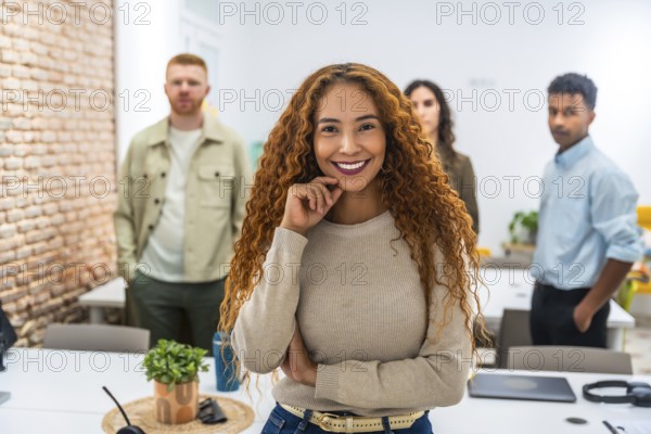 Confident young businesswoman with curly hair smiling at camera. Standing in a contemporary office space while her diverse colleagues stand blurred in the background. Representing teamwork and success