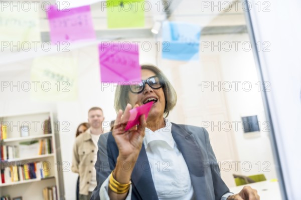 Mature businesswoman attaching colorful sticky notes on a clear glass board, collaborating on strategic ideas during a business meeting with colleagues in the modern office