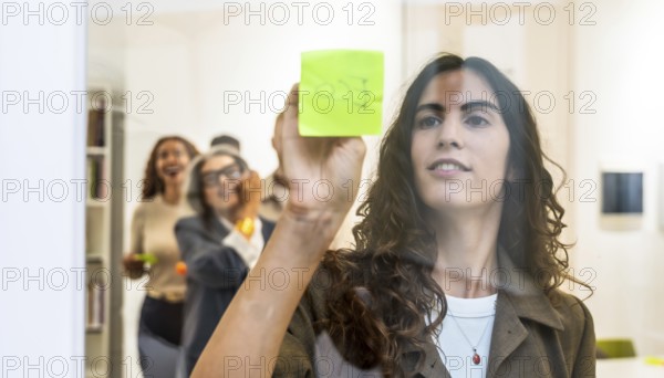 Young businesswoman sticking a green sticky note on a transparent glass wall during a team meeting, her colleagues in the background engaging in collaborative brainstorming and creative discussions