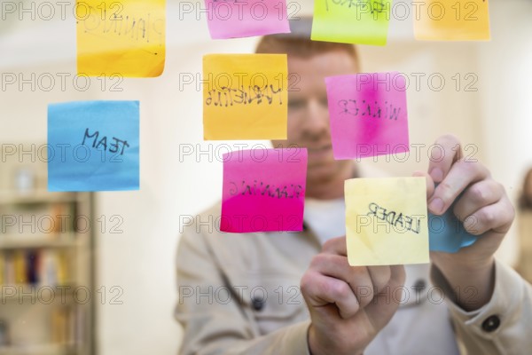 Red haired man organizing colorful sticky notes with handwritten words about planning, team, and leadership on a clear glass board during a focused brainstorming session at the office