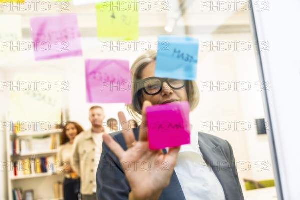 Mature businesswoman arranging colorful sticky notes on a glass board while leading a strategic planning session with her team in a modern office, focused on collaboration and goals