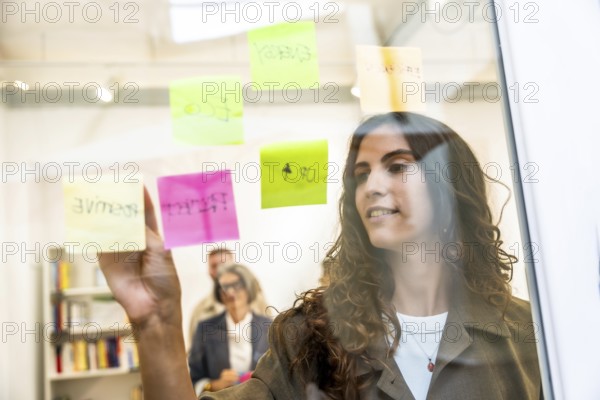 Young professional woman brainstorming and planning new business ideas, organizing strategy, and writing notes on a transparent glass wall in a modern office while colleagues work in the background