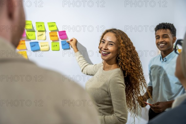 Young diverse business professionals collaborating, smiling, and brainstorming creative solutions during a teamwork meeting, sticking colorful notes on a whiteboard in a modern office setup