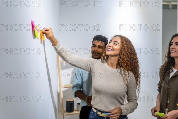 Young diverse team collaborating on a project, happy woman placing a colorful sticky note on a white wall during a brainstorming session in a modern office