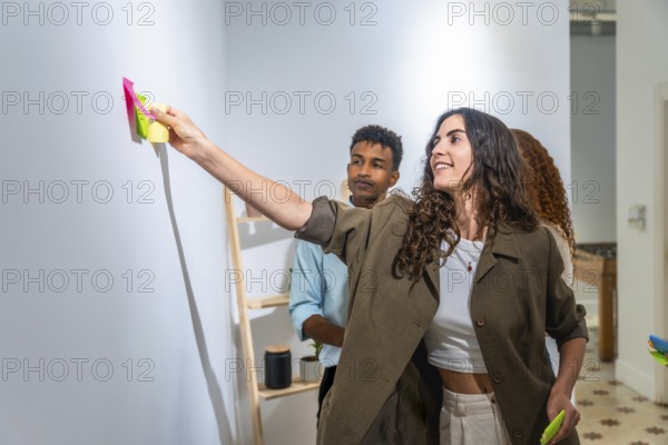 Diverse team collaborating, standing in a modern office, a smiling woman adding colorful sticky notes to a white wall during a creative brainstorming session