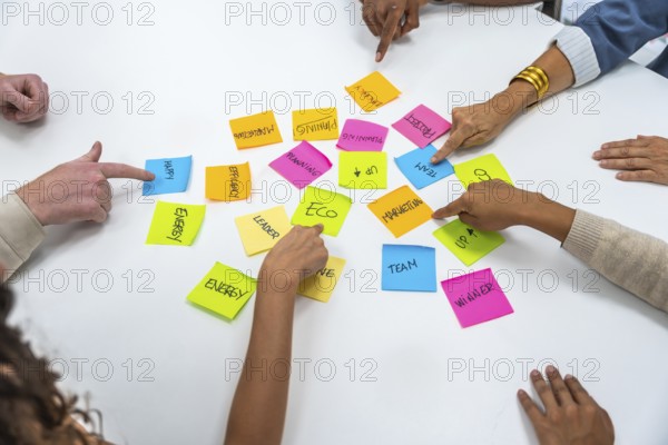 Diverse business professionals collaborating, pointing at colorful sticky notes with written concepts on a white table, symbolizing teamwork, planning, and strategy in a corporate meeting setting