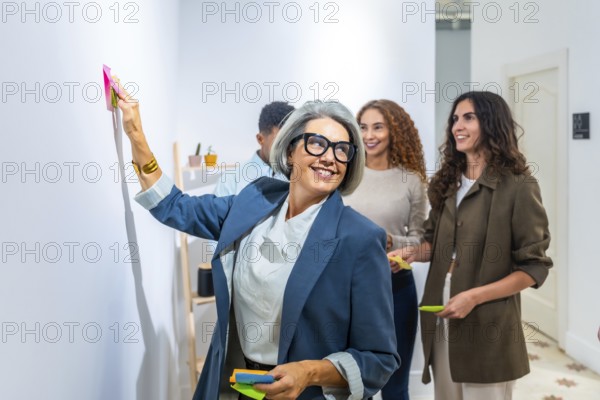 Confident gray haired businesswoman in glasses posts a sticky note during a lively team brainstorming session, leading collaboration and innovation in a modern office setting